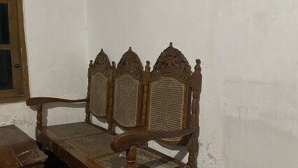 a long wooden chair with a classic design placed in a room with dull white walls. This chair has a unique backrest with distinctive carvings and woven rattan in the middle