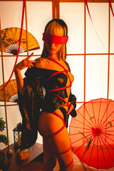 Beautiful young woman with blonde hair, black kimono and panties, and red shibari ties and ropes, red blindfold, holding a paper fan in a warm japanese room