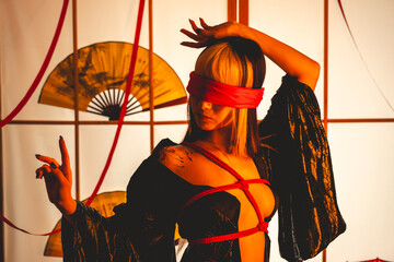 Obraz premium Beautiful young woman with blonde hair, black kimono and panties, and red shibari ties and ropes, red blindfold, holding a paper fan in a warm japanese room