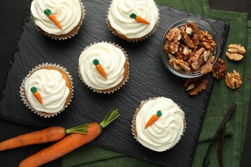 Tasty carrot muffins with cream, walnuts and fresh vegetables on black wooden table, flat lay