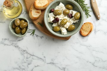 Marinated olives with feta cheese, bread pieces, oil and rosemary on white marble table, flat lay. Space for text