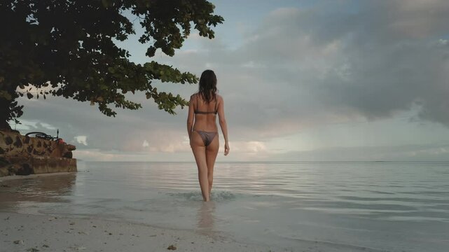 Young woman in a bikini is enjoying a relaxing evening on a tropical beach, walking into the calm ocean water as the sun sets over the horizon