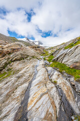 A stunning view of the Schlaten Glacier area in Austria, featuring rugged, layered rocky terrain illuminated by soft sunlight under a partly cloudy sky. The landscape showcases the beauty of Alps