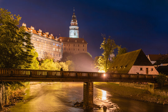 The Cesky Krumlov Castle Tower stands majestically illuminated against the night sky. Its reflection glimmers on the river below, creating a stunning backdrop for visitors.