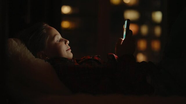 Smiling 10 yo child girl lying comfortably on bed, using smartphone in dark bedroom at night, with only bright screen illuminating face, creating cozy and relaxed atmosphere. Shooting in slow motion.
