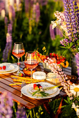 Romantic table decor for a loving couple on blooming meadow with purple lupines. Two glasses of wine, flowers in a vase, silverware, fruits, wooden furniture. Sunset, golden hour, atmospheric light