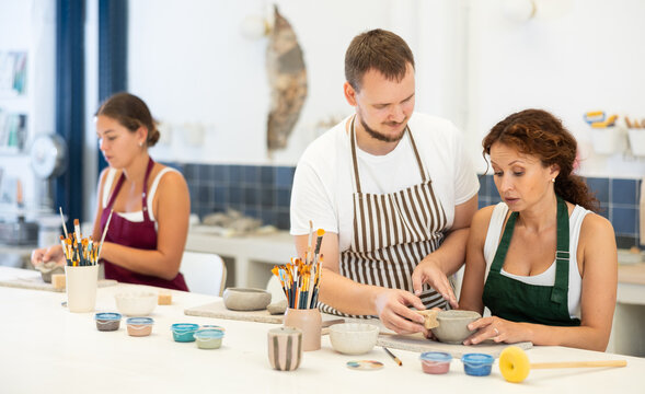 In pottery workshop, apprentice female students sitting at table, engaged in making products from raw clay in presence of teacher. Visitors to clay craft workshop work under guidance of male mentor - Powered by Adobe