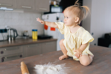 Playful toddler girl sitting amid scattered flour, reaching hands across messy kitchen table,...