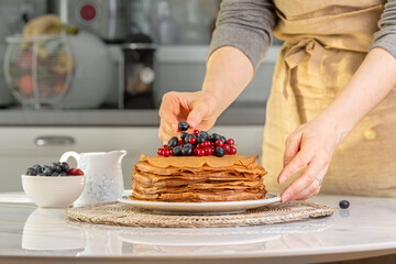 Woman in apron putting berries on a stack of sweet homemade crepes, type of thin pancake, with blueberry and red currant in the center of table.