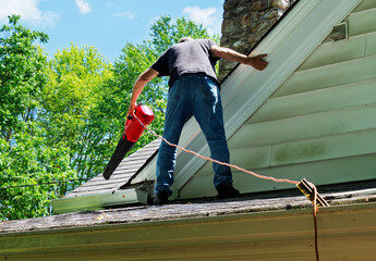 Man standing on porch roof to clean a gutter with leaf blower