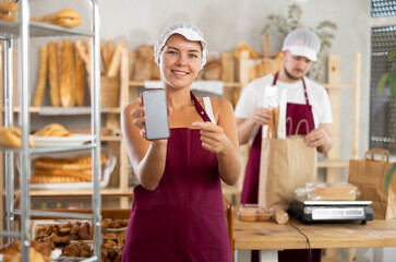 Positive young female worker displays a mobile application while male seller is packing baguettes...