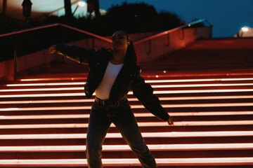 Confident Hispanic woman enjoying music in wireless earbuds while dancing on illuminated city stairs at night. Dressed in leather jacket and jeans,she expresses freedom,joy, and modern urban lifestyle