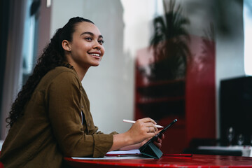 Smiling Latina entrepreneur working remotely from trendy café,using digital tablet and stylus for...