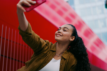 Smiling young Latina woman taking selfie outdoors. Female enjoys capturing moments, reflecting...