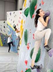 Woman is mastering climbing on training wall in gym. Woman holds on tightly to ledges and strives...