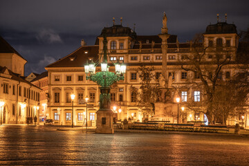 In the evening, the historic lamppost at Hradcanske Square casts a warm glow over the cobblestone streets. Surrounding buildings reflect light, enhancing the lively atmosphere of Prague.