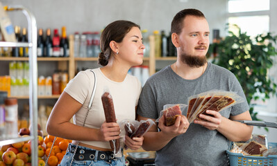 Portrait of satisfied male and female customers with dried sausage and jamon in hands shopping together in grocery store