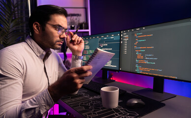 Smart IT developer working on software development coding on pc screen and reading note of newest application project, thinking program update online website data at neon light cyber office. Surmise.