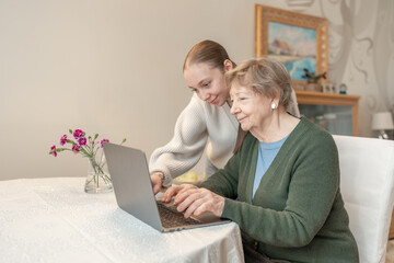 Young woman helping a senior woman with using laptop at home. Concept of technology, education, and...