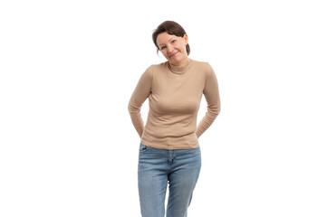 Fototapeta premium A young woman in a beige long-sleeve top smiling and standing with her hands behind her back against a white background.