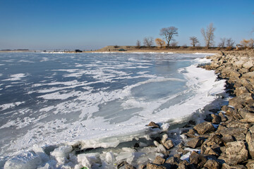 Frozen lake surface with snow and rocks on the shore and bare trees in the background during a sunny winter day