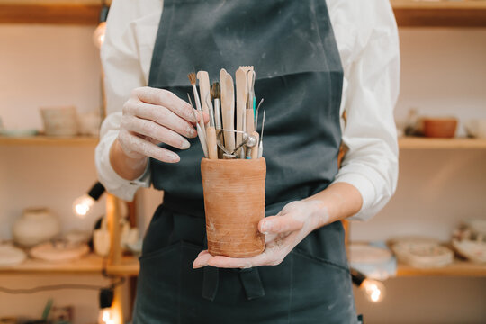 Clay crafting tools for ceramists. Potter in black apron holds a bunch of instruments for sculpting pottery in ceramic mug.