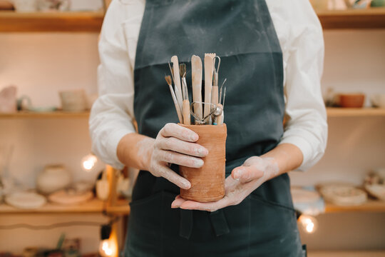 Clay crafting tools for ceramists. Potter in black apron holds a bunch of instruments for sculpting pottery in ceramic mug.