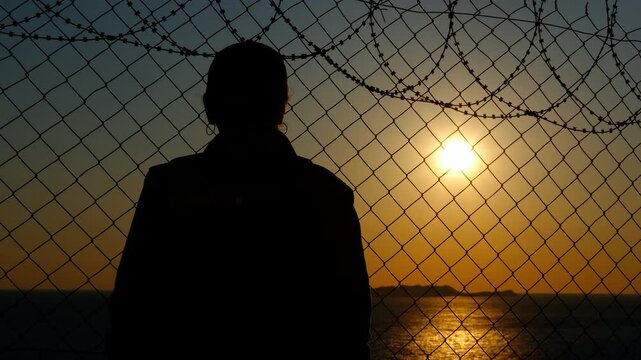 A refugee in despair at the border. A silhouette of a refugee desperately clings to the barbed wire at the border against the backdrop of a sea sunset