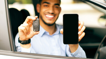 Cheerful middle-eastern businessman sitting inside nice auto, showing car key and smartphone with blank screen, positive arab man recommending online service for car renting, mockup, copy space