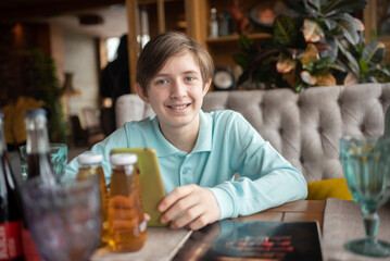 Smiling boy holding mobile phone, sitting at table in cafe, waiting for his order in restaurant.