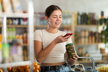 Young woman buyer scanning qr code for pickled cucumbers in jar in grocery store