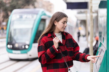 Girl passenger purchases reusable travel pass from machine at tramstop. She types desired number of trips on screen and selects ticket price.