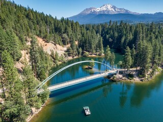 Aerial view of a pedestrian bridge over a serene Lake Siskiyou, surrounded by lush forest and a Mount Shasta snow-capped mountain. A small boat floats on the water. California, USA