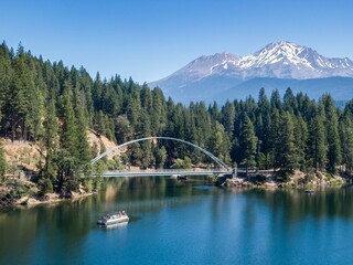 People enjoy a boat ride on a beautiful lake, beneath Mount Shasta mountain range. A scenic bridge...