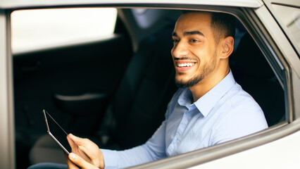 Cheerful middle-eastern businessman checking emails while going to airport by taxi, sitting on car back seat and using digital tablet, closeup photo, shot from outside, side view, copy space