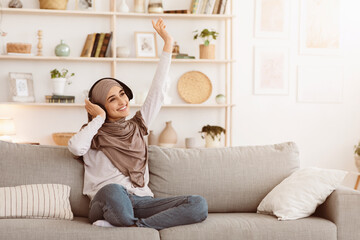 Obraz premium Cheerful Day. Positive Arabic Girl In Hijab Listening Music In Wireless Headphones At Home, Relaxing On Comfortable Couch, Copy Space