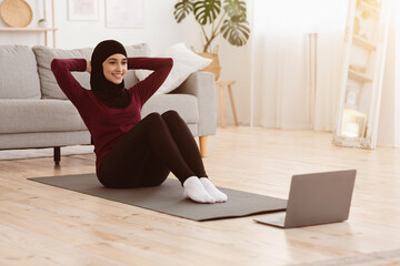 Fit arabic woman training at home, doing abs exercises while watching tutorial videos on laptop,...