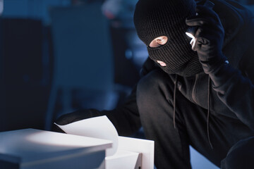 Closeup of disguised robber with flashlight searching for documents in office at night, copy space, blurred background