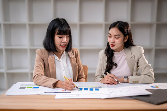 Two women are sitting at a desk, one of them writing with a pen