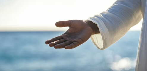 Close-up of Jesus's hand reaching out to help, white sleeve, blurred background of sky and beach Generative AI