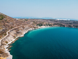 Obraz premium Aerial view showcasing a winding coastal road along a rocky shoreline, with vibrant turquoise waters and distant mountains under a clear blue sky in Ko Sichang, Chonburi, Thailand