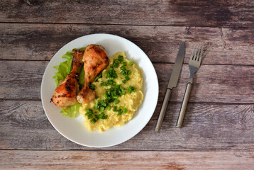 Two baked chicken legs on a plate with mashed potatoes and green onions on a wooden table.