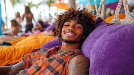 Fototapeta premium A young man with curly hair smiles while lounging on a bright purple chair at the beach. Friends can be seen enjoying the sunny day around him, creating a lively atmosphere
