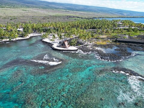An aerial view of the rugged lava rock shoreline of Pu'uhonua o Honaunau, with the vibrant turquoise ocean surrounding it and lush green mountains in the distance. The natural landscape offers a drama