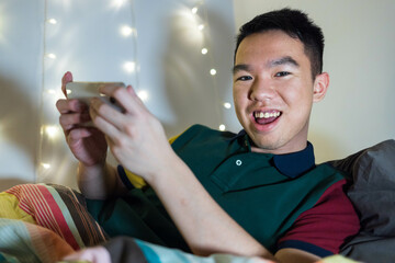 A young Asian male teenager is using his phone to play video games on his bed at home. He is very...