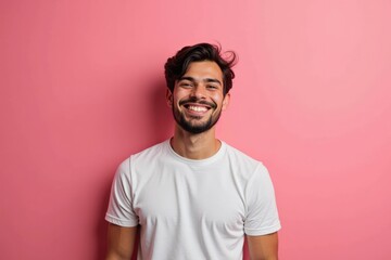 Obraz premium Smiling Young Adult Male With Dark Hair Standing Against a Pink Background, Exuding Happiness and Confidence in a Casual White T-Shirt