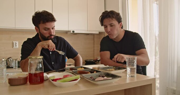 Two Caucasian men enjoy a healthy breakfast together in their modern kitchen. They talk and laugh while eating eggs, vegetables, and fruit.