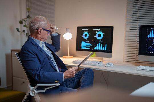 Stressed senior businessman having headache while working late at night in office analyzing financial data on computer and tablet, holding pen in hand and touching his forehead