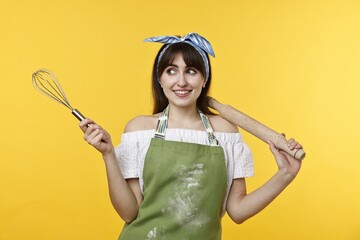 Happy woman with rolling pin and whisk on yellow background