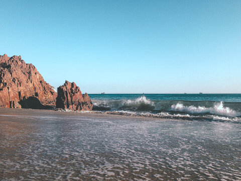 playa con arena y piedras rocosas grandes con olas en una tarde soleada y cielo azulado
